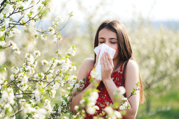 Allergy season, young woman blowing her nose and sneezing in blossoming garden.